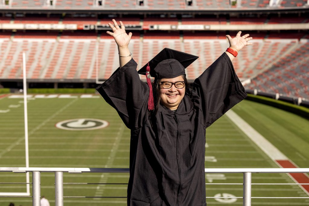 Marina Martinez wears a graduation cap and gown throws her hands up in excitement. Football field in the background.