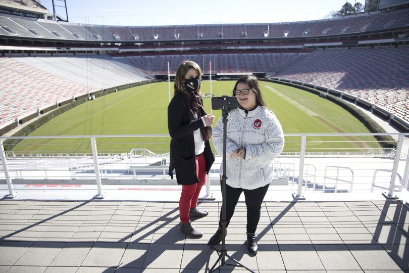 Whitney Ostrander and Marina Martinez stand in front of a football field and film on an iPhone.