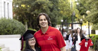 Marina Martinez wears a graduation gown and poses for a photo with her brother in front of UGA's Arch.