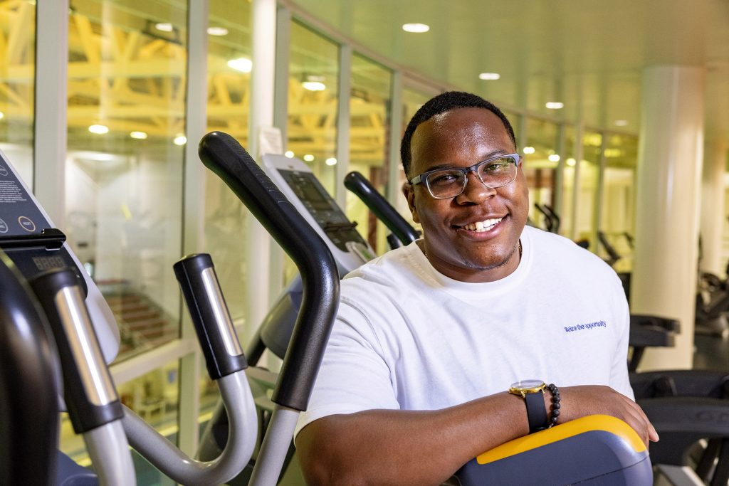 Zerian Hood smiles as he stands in a gym full of exercise equipment