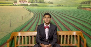Jay Ivey, wearing a suit and bow tie, sits on a bench in front of a mural of farmland.