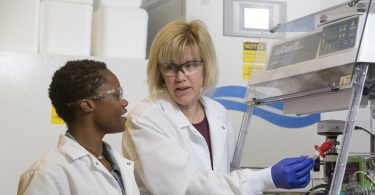 Karen Burg (right) talks to Kristen Allen (left). Both women are wearing white lab coats and safety glasses.