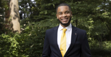 Portrait of Eric Okanume outdoors. He is wearing a suit with a yellow tie.