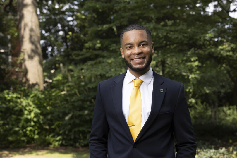 Portrait of Eric Okanume outdoors. He is wearing a suit with a yellow tie.