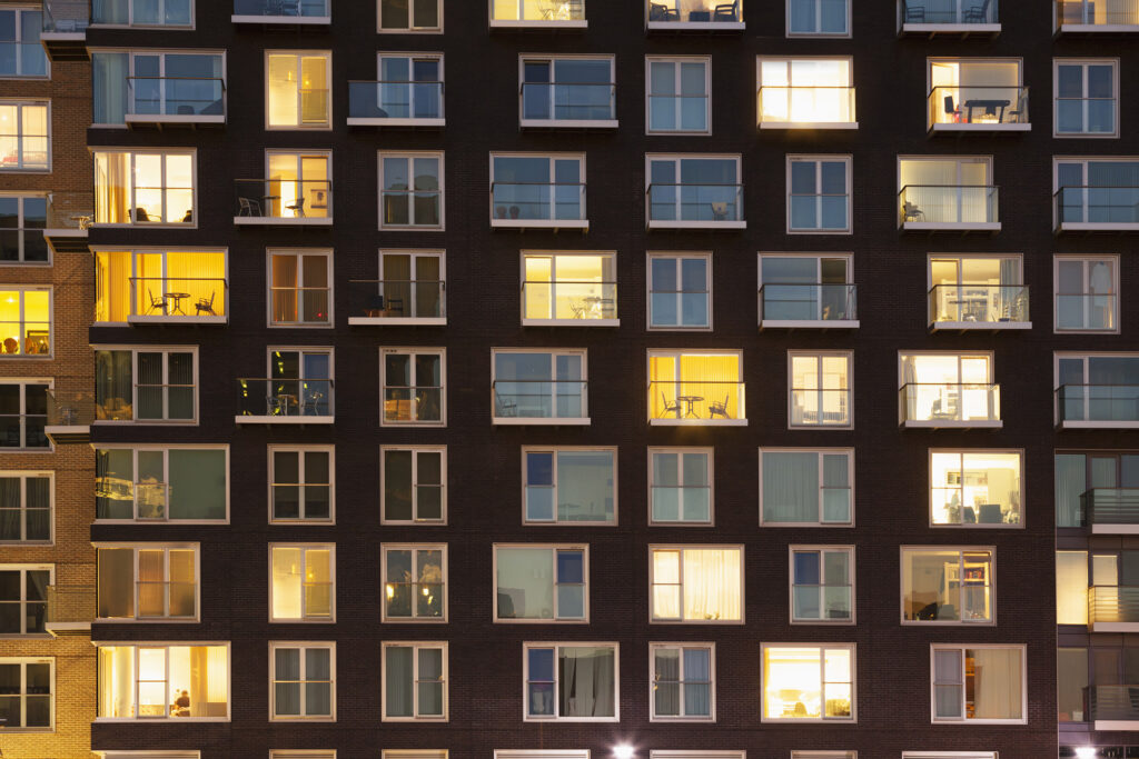 People are shown in the windows of their units in a large apartment building at dusk.