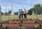 Three people stand behind newly planted raised garden beds.