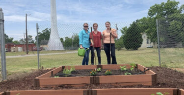 Three people stand behind newly planted raised garden beds.
