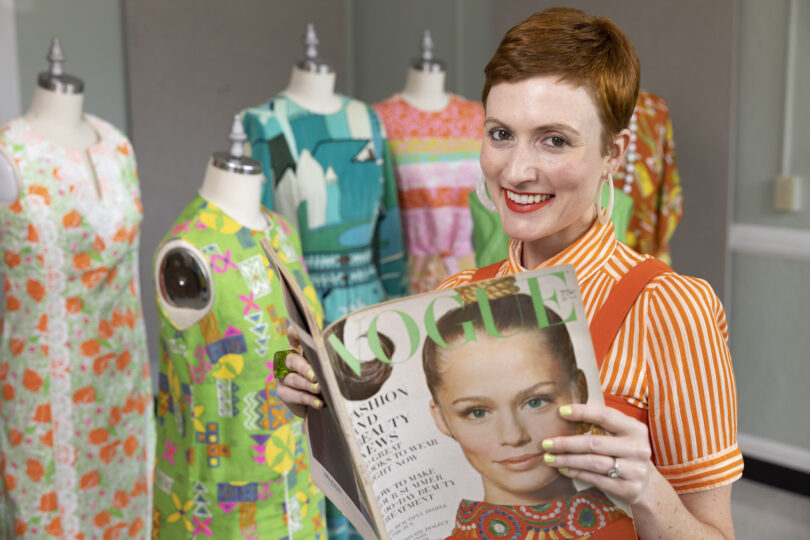 Sara Idacavage holds a vintage magazine in front of vintage clothes.