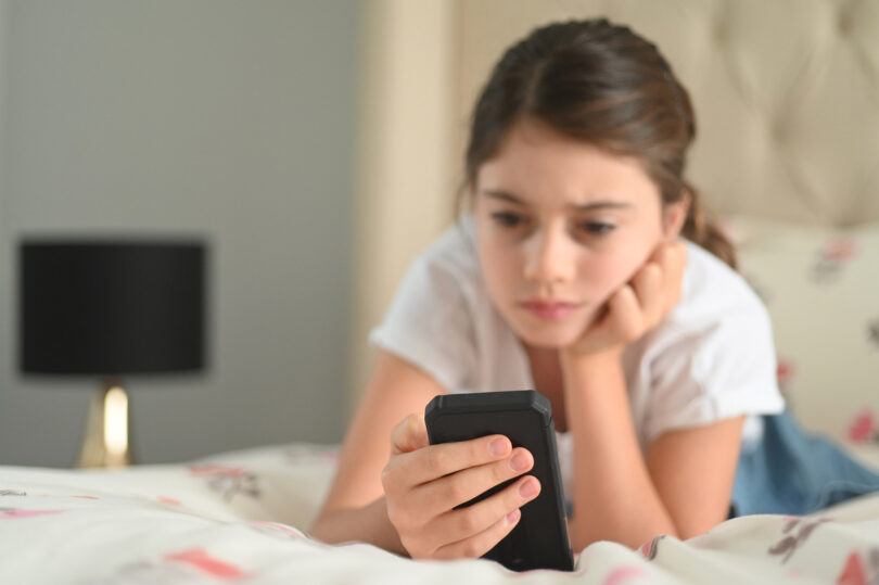 A young girl looks at her phone with a sad expression on her face.