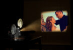 A film projector projects a scene from a movie on a screen. The image shows a couple.