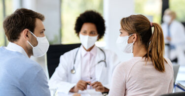 Young couple wearing protective face masks and holding hands while having appointment with a doctor at medical clinic.
