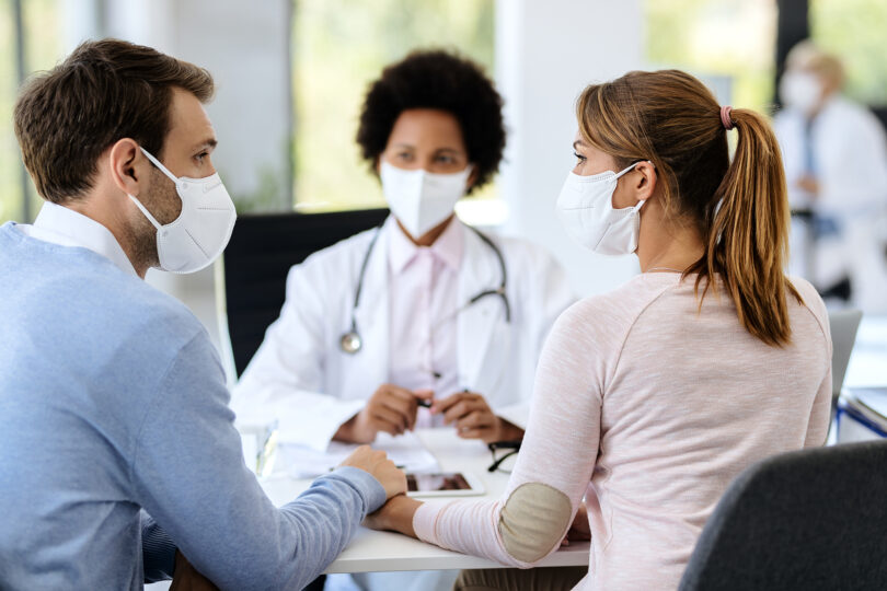 Young couple wearing protective face masks and holding hands while having appointment with a doctor at medical clinic.
