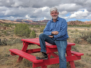 R. Phillip Bouchard is sitting on top of a red picnic table with his arms crossed, smiling at the camera. A hilly desert landscape is seen behind him.