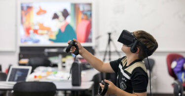 A child wearing a virtual reality headset plays a game in a classroom setting.