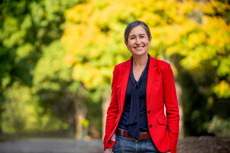 Female researcher stands outside in a red jacket smiling. Her research is about the secret to happiness.