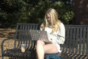 Mikaela Haast sits outside on a sunny day on UGA's north campus working on her computer. Her computer has a case covered in white starts, and a sticker of the Tri Delta sorority letters. 