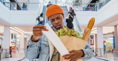 A Black man in a jean jacket and orange beanie looks at his grocery receipt because he is concerned with inflation