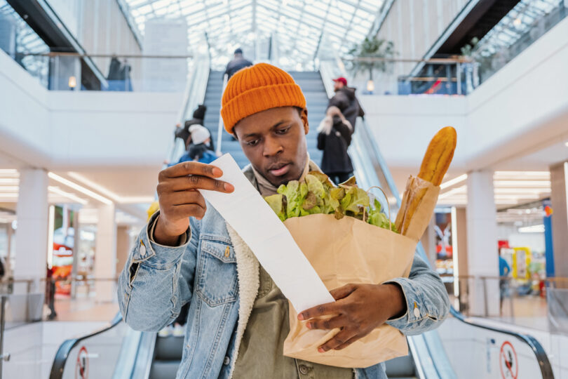 A Black man in a jean jacket and orange beanie looks at his grocery receipt because he is concerned with inflation