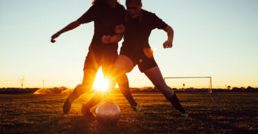 Two soccer players work on their skills with drills at dusk with the sun shining through their silhouettes.