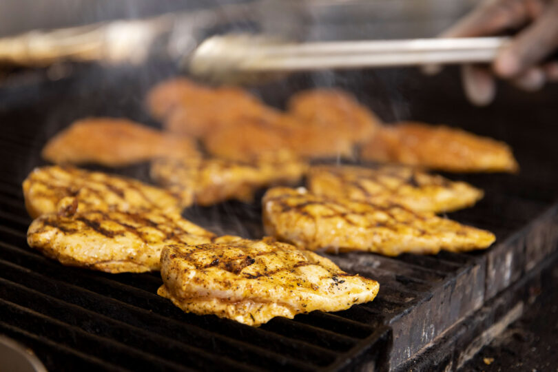 Close up of chicken cooking on a grill with a hand holding tongs in the background.