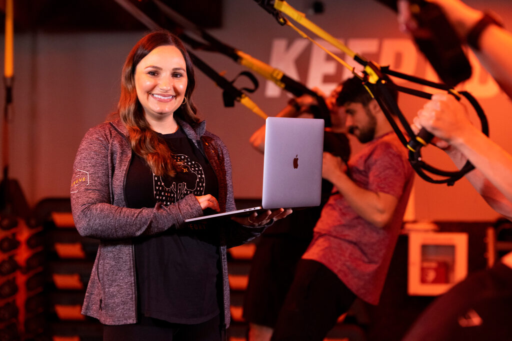 Rachelle Reed is pictured inside of an Orange Theory gym, holding her laptop and smiling at the camera.