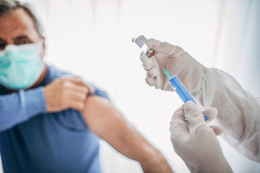 An older man rolls up his sleeve while wearing a mask to receive a COVID-19 vaccine from a health care worker who is wearing gloves.