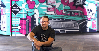 Marc Gorlin is pictured sitting on the ground in front of a blue and pink mural.