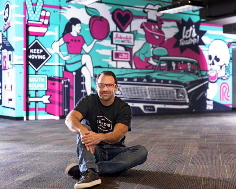 Marc Gorlin is pictured sitting on the ground in front of a blue and pink mural.