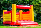 A red and yellow bounce house, or bouncy castle, sits on a lawn with trees in the background on what appears to be a sunny day.