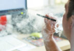 A man is shown from the side smoking an e-cigarette in front of part of a computer screen.