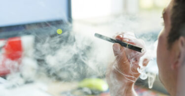 A man is shown from the side smoking an e-cigarette in front of part of a computer screen.