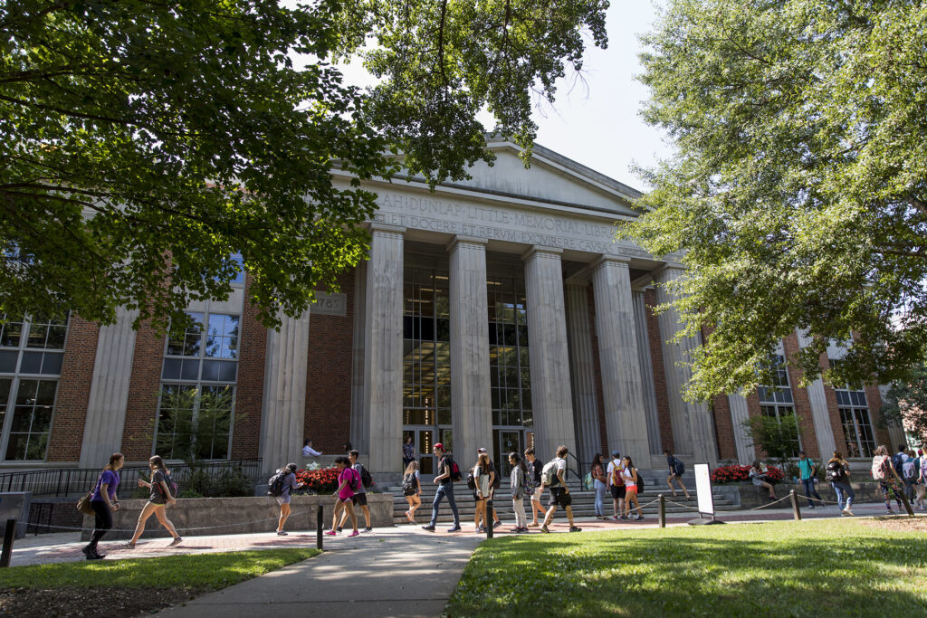 Students walk on a pathway through green lawns. In the back ground is a majestic library of red brick with off-white pillars