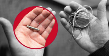 Two hands. One is holding a small bullet shaped device that is a wireless pacemaker. The right hand holds an older model pacemaker, a square with several leads sticking out.
