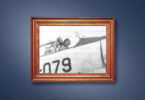 A wooden picture frame on a blue wall holds a grainy black and white photo of pilot in a jet.