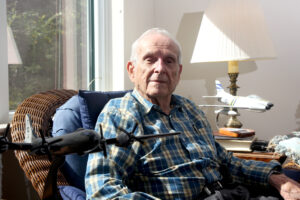 A man in his 90s sits in a wicker chair with two model aircraft (a bomber and a fighter jet) sit on either side of him.