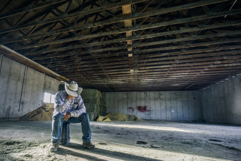 Inside a barn, a desperate looking man sitting all by himself on a bucket.