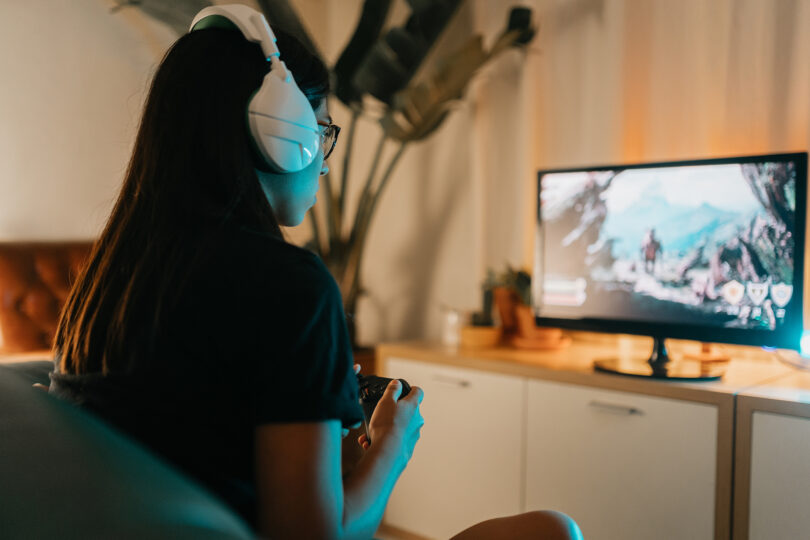 A young woman with long black hair wearing headphones plays a video game.
