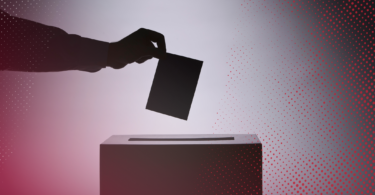 A silhouetted hand places a ballot into a ballot box.