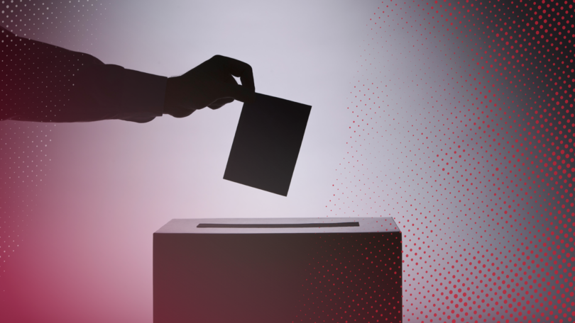 A silhouetted hand places a ballot into a ballot box.
