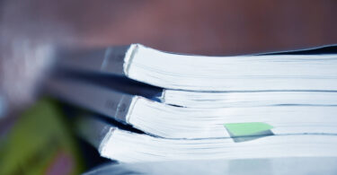 A stack of scientific journals are shown stacked on a desk with a sticky note on one of them.