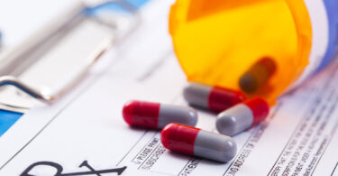 Red and blue pills are shown spilling out of an orange prescription bottle onto a prescription pad.