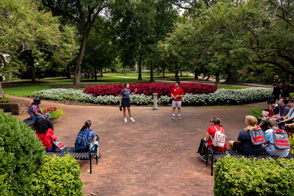 Two UGA Visitors Center tour guides stand in front of a group of people touring campus.