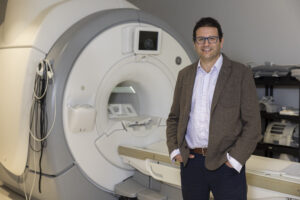 Environmental portrait of Assaf Oshri in front of an MRI scanner used for his research.