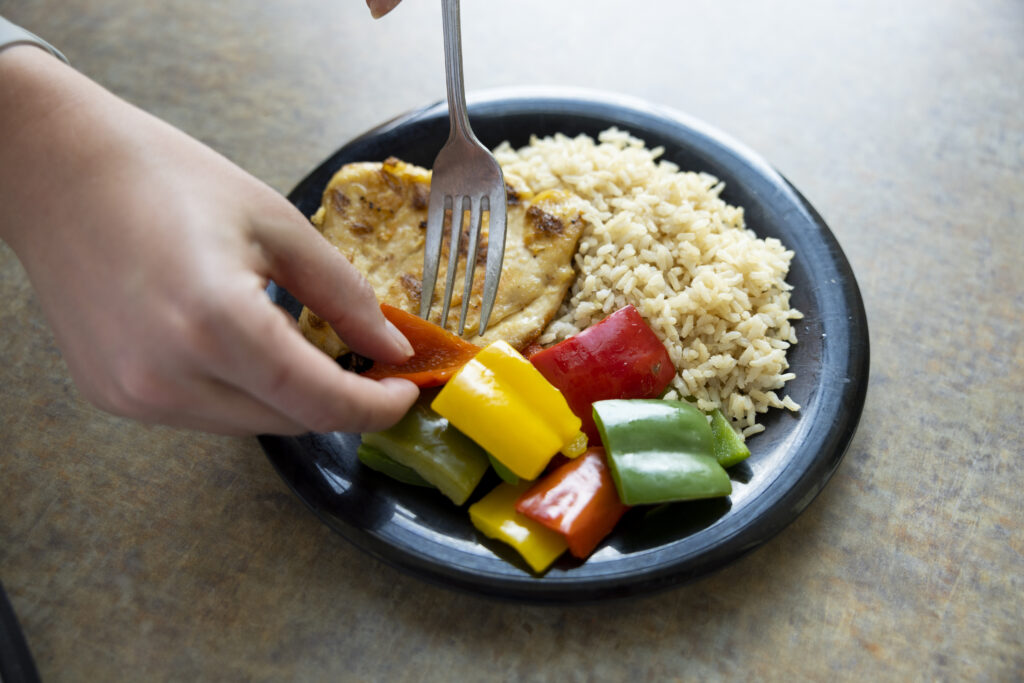 A hand is shown with a fork eating a plate of chicken, rice and cooked peppers.