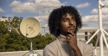 Nathan Tesfayi poses with one of the communications antennas used by the small satellite lab on the roof of the Geography Geology building.