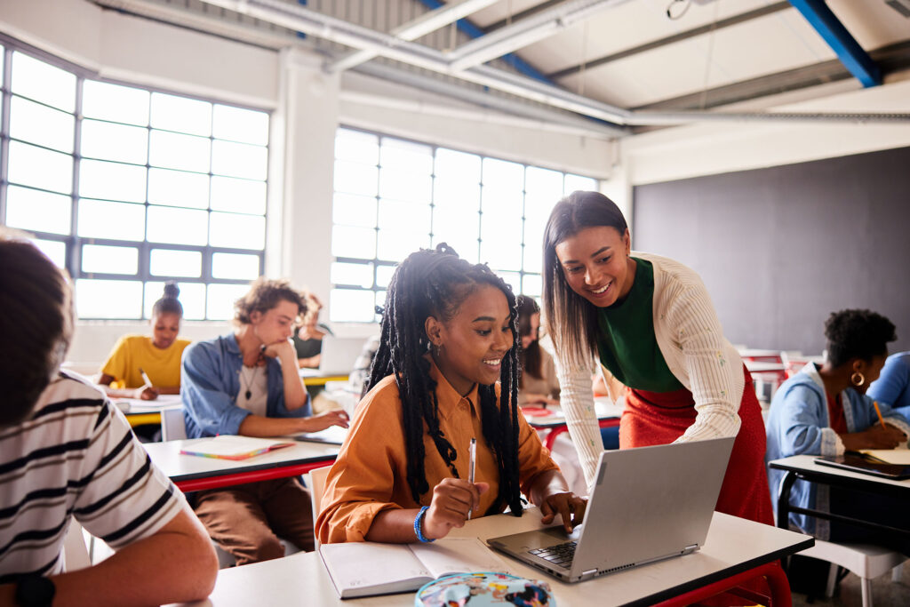 Two women, one seated and one standing, hover over an open laptop in a sunlit classroom with another male student in the background.