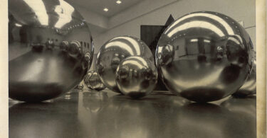 In this black and white photo, large, reflective shiny metal ball sculptures stand in an art gallery