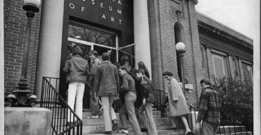 In black and white, a group of high school age students walk up the steps into a building with a sign that reads Georgia Museum of Art.
