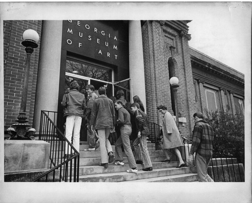 In black and white, a group of high school age students walk up the steps into a building with a sign that reads Georgia Museum of Art.