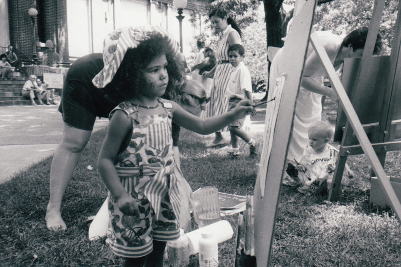 In this black and white photo, young girl, maybe 4 years old, paints a picture on an easel while standing in the grass.